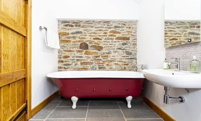 A bathroom with a red roll-top bathtub, white sink, slate floor tiles, and a rustic exposed stone wall, with a wooden door and modern fittings